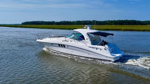 White motorboat speeding through calm water with marshy grass and a tree-lined shore in the distance.