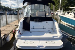 View from the stern of a white boat at a marina, with a black canvas cabin and other docked boats along the pier behind blurred water reflections.