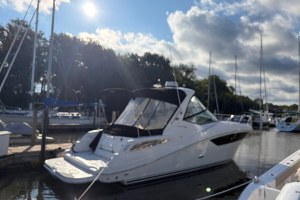 Yacht docked at a marina with tall masts, calm water, and a sunny, partly cloudy sky behind trees in the background.