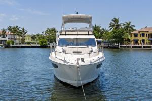 Front view of a white motor yacht moored in a calm harbor with palm trees and seaside houses in the background.