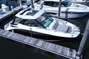 White powerboat docked at a marina with cushioned seating and a glass windshield, tied to a wooden pier with a larger yacht in the background.