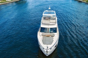 Top-down view of a white motor yacht cruising on blue water, with a dock visible along the shore on either side.