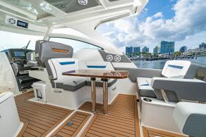 Modern yacht cabin lounge with white and gray seating around a polished wooden table on a teak deck, marina and skyline in the background.